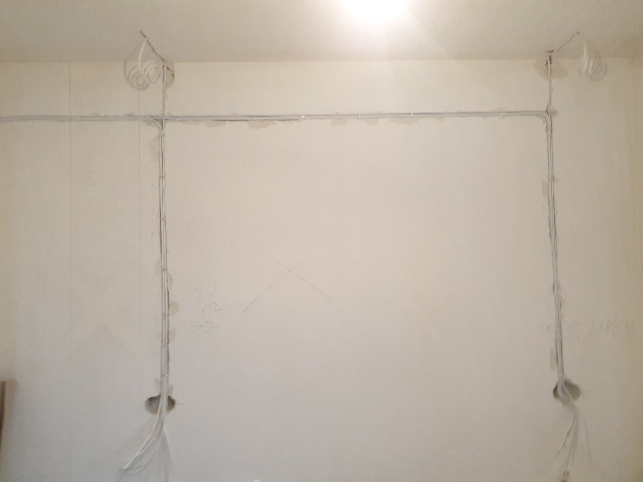 Szare kable elektryczne ułożone wzdłuż ściany i sufitu w jasnym pomieszczeniu, widoczne otwory w ścianie i kable zwisające z sufitu.