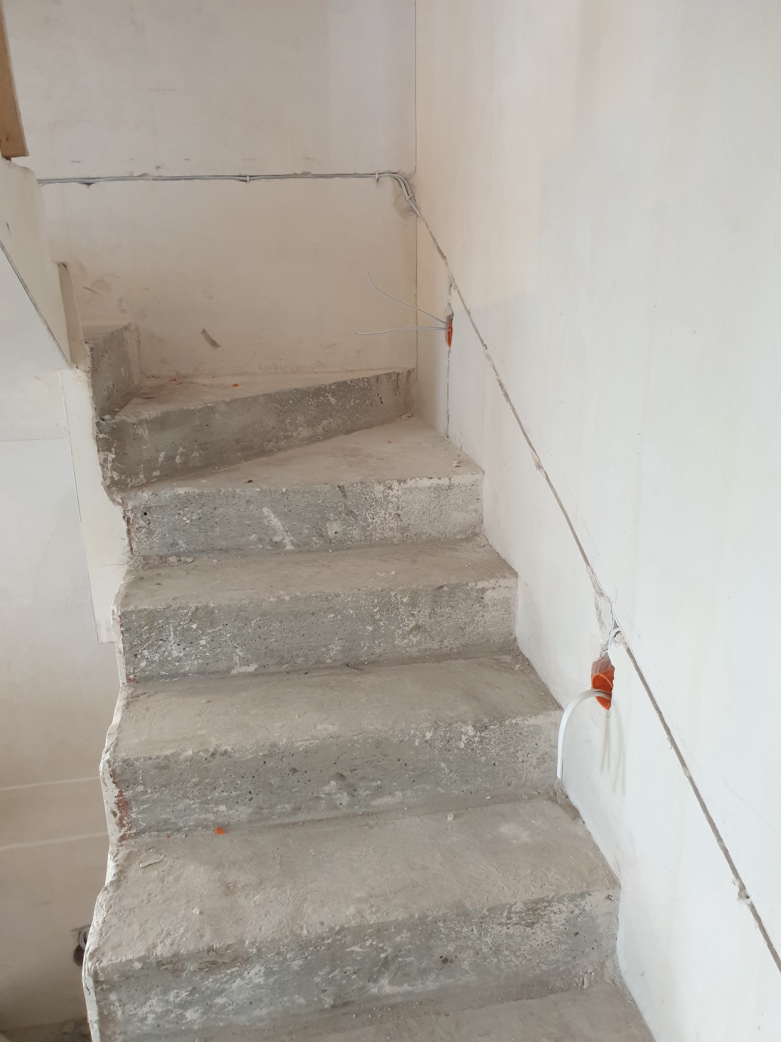 Surowe betonowe schody w trakcie remontu, z widocznymi przewodami elektrycznymi wystającymi ze ściany i z puszek instalacyjnych, szare przewody ułożone wzdłuż ściany.
