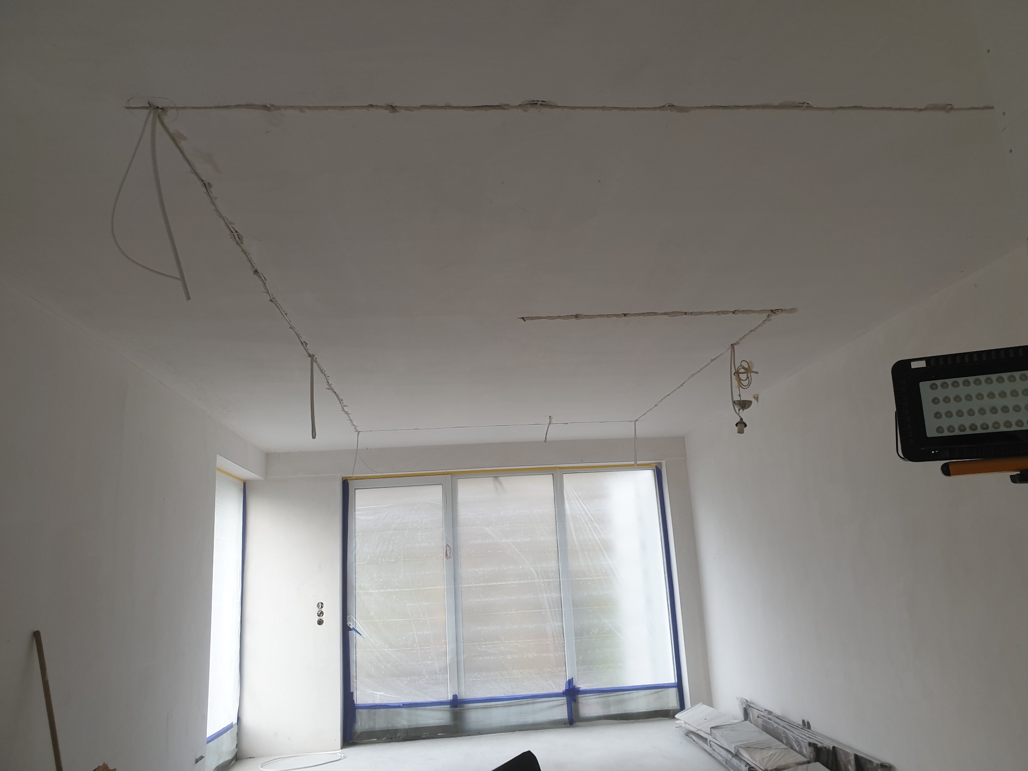 Wnętrze w trakcie instalacji elektrycznej: widoczne przewody w suficie, przygotowane punkty oświetleniowe oraz zabezpieczone okna folią malarską.