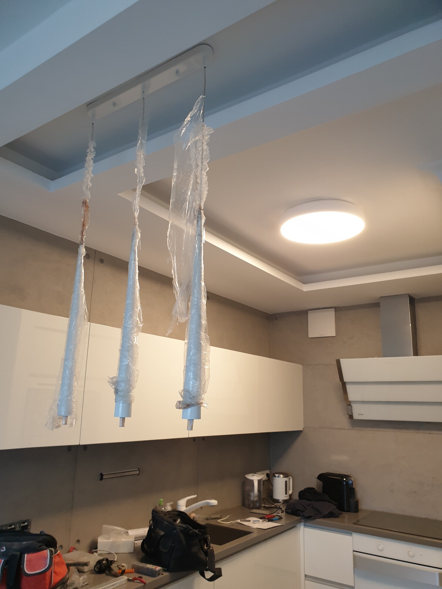 Nowoczesna lampa wisząca z trzema kloszami w folii ochronnej, zamontowana w kuchni z białymi szafkami i szarymi ścianami, widoczna okrągła lampa sufitowa.