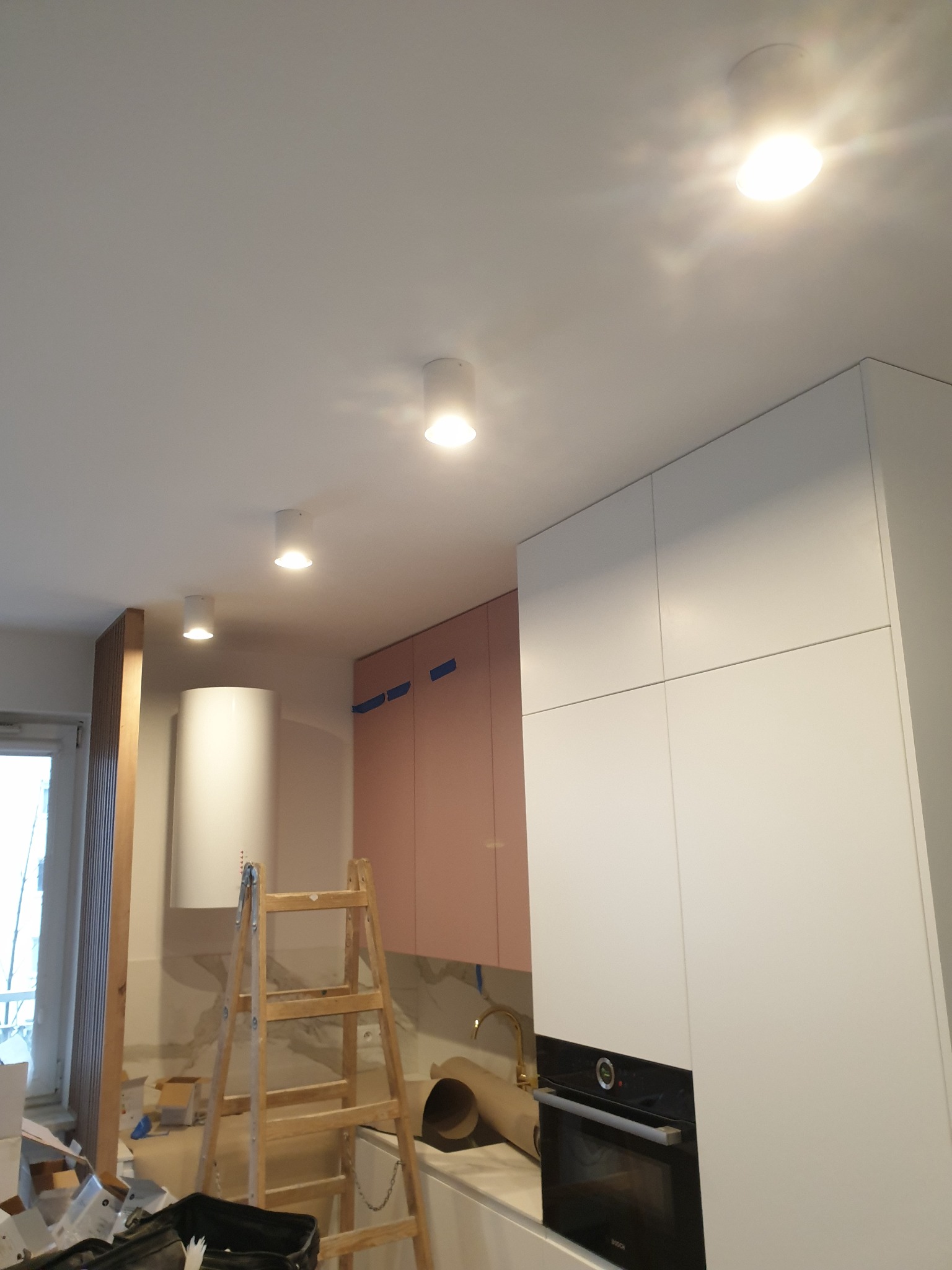 Białe, okrągłe lampy sufitowe zamontowane w kuchni w trakcie remontu, widoczna drabina i częściowo zamontowane meble kuchenne.