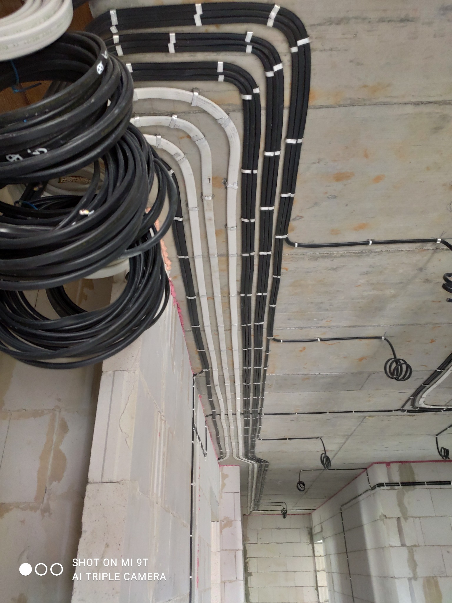 Wiązka czarnych kabli zwisająca ze ściany z betonu komórkowego, równolegle ułożone peszle z przewodami elektrycznymi, przytwierdzone metalowymi uchwytami do sufitu z betonu architektonicznego...