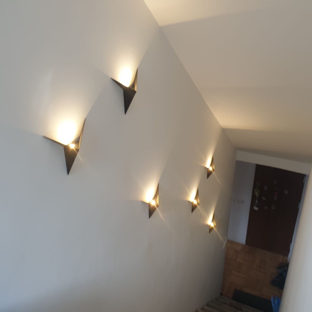Rząd designerskich kinkietów w kształcie strzałek, oświetlających schody w korytarzu, z widoczną fakturą ściany.