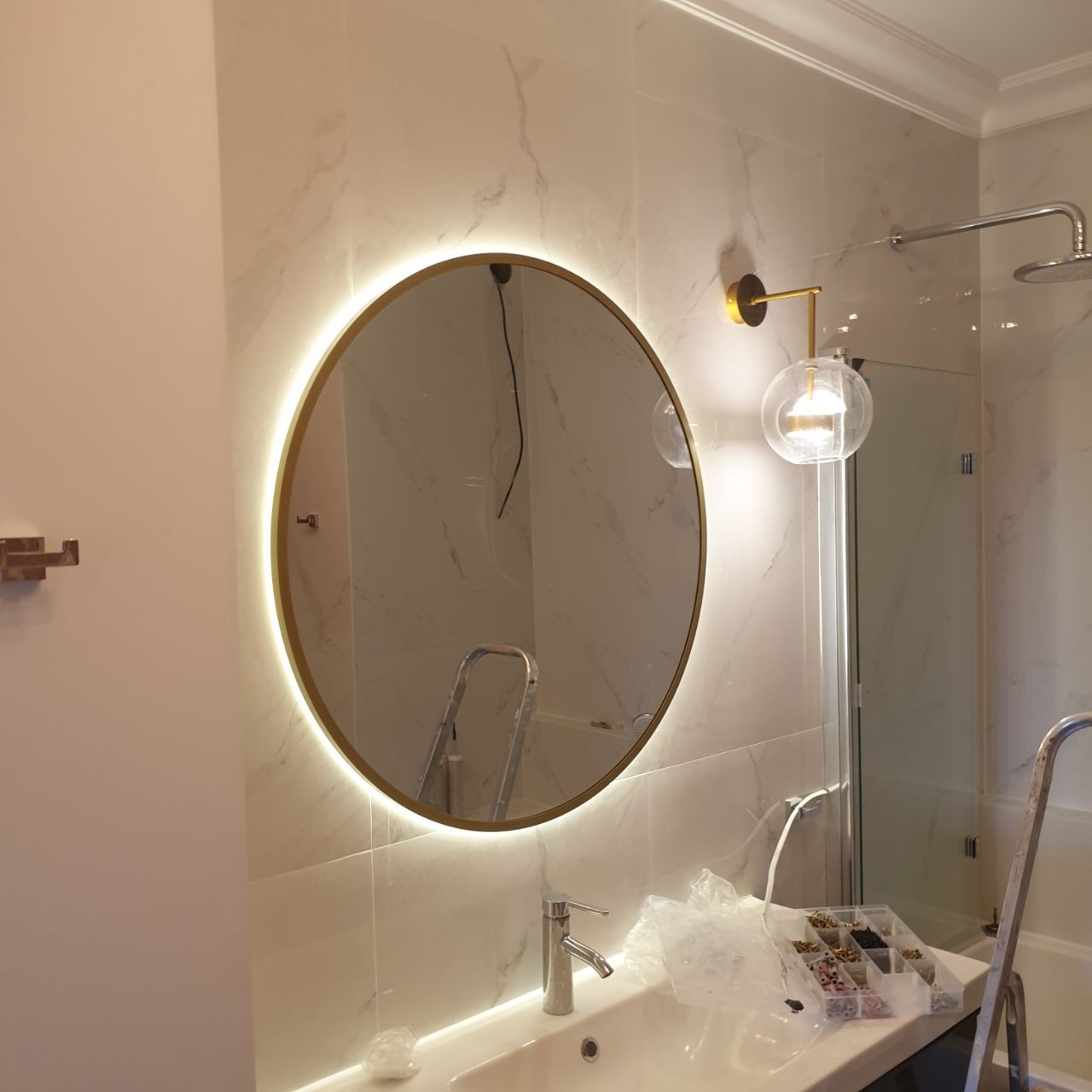Okrągłe lustro z podświetleniem LED w łazience z marmurowymi płytkami, widoczna szklana lampa ścienna, drabina i akcesoria do montażu na blacie umywalki.