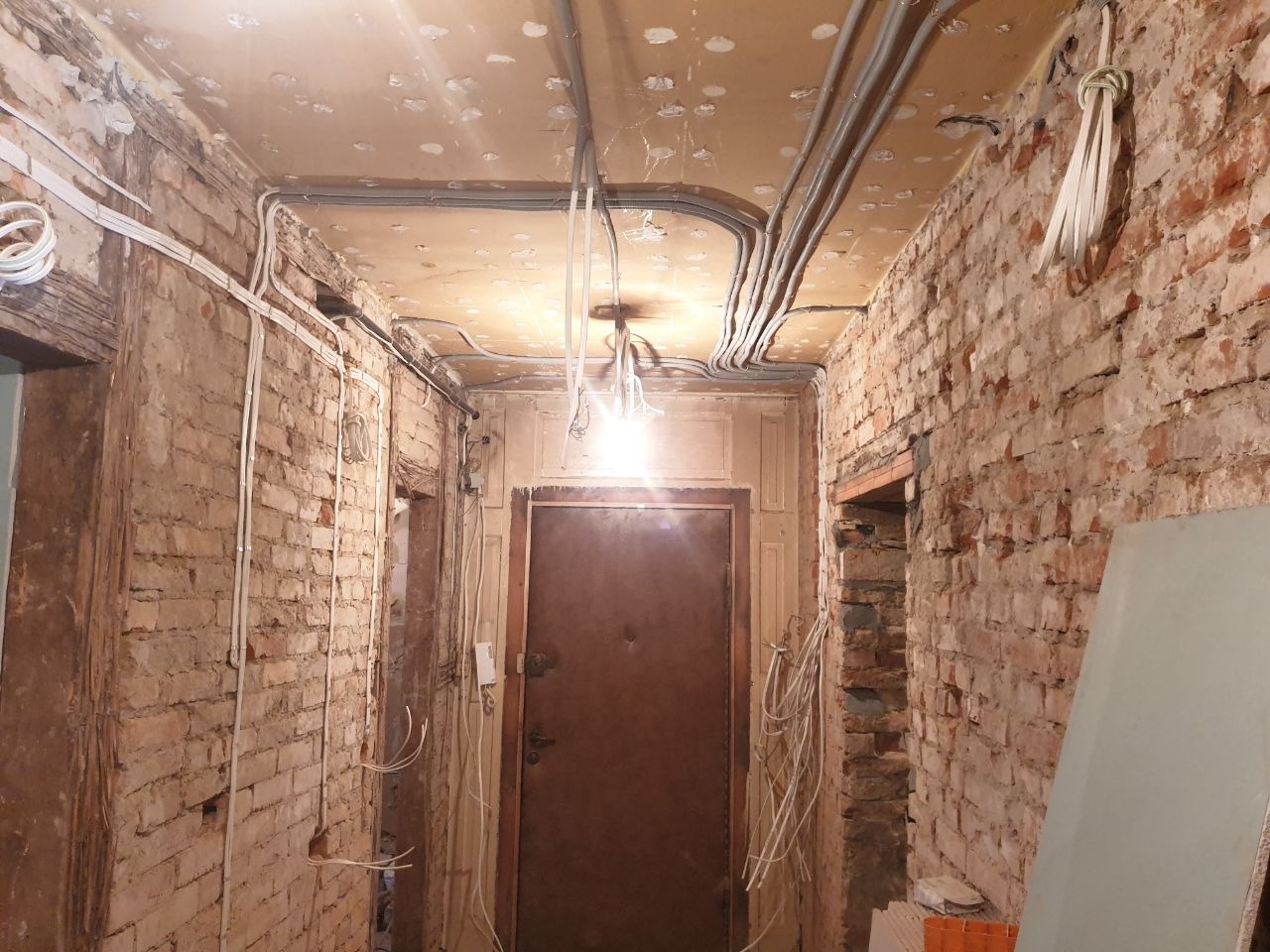 Korytarz w trakcie remontu z odsłoniętą cegłą, widoczne szare kable elektryczne ułożone na suficie i białe kable biegnące po ścianach, zwisające kable ze środka sufitu.