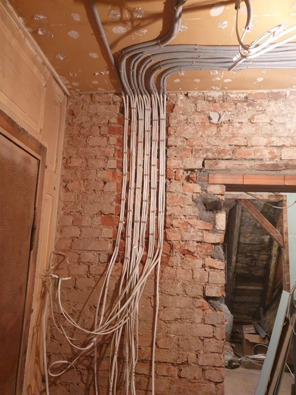 Równoległe wiązki przewodów elektrycznych w białej izolacji, prowadzone po ścianie z czerwonej cegły w trakcie remontu, z widocznymi elementami instalacji w peszlu pod sufitem.