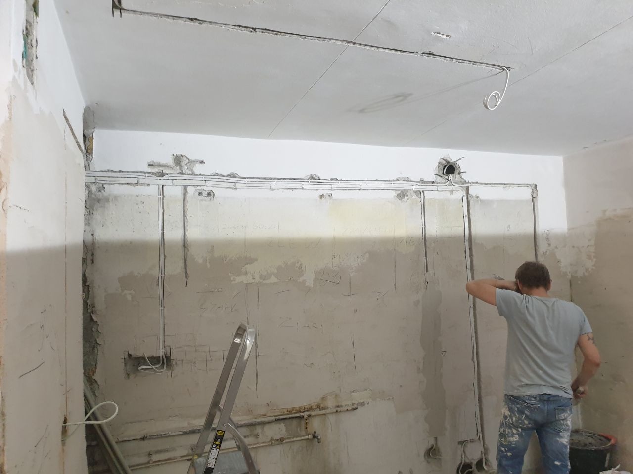 Elektryk w trakcie instalacji elektrycznej w pomieszczeniu w stanie surowym; widoczne kable prowadzone po ścianach, drabina aluminiowa, tynk w trakcie kucia
