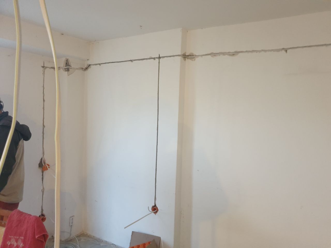 Przewody elektryczne prowadzone po ścianie w trakcie prac instalacyjnych, widoczne puszki instalacyjne i fragment osoby w odzieży roboczej.