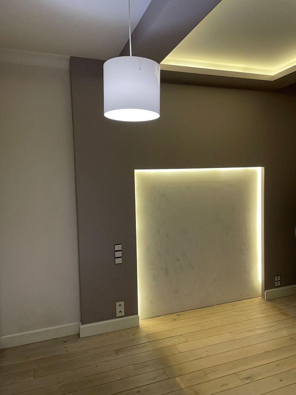 Nowoczesny salon z podświetlaną wnęką w ścianie, wiszącą białą lampą cylindryczną i drewnianą podłogą w jodełkę, widoczne gniazdka i włączniki.