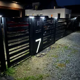 Nowoczesna, czarna brama wjazdowa z numerem domu '7' podświetlonym od wewnątrz, oświetlenie bramy LED, szara nawierzchnia z kamienia na podjeździe, widok na oświetlony dom w tle, nocne ujęcie.