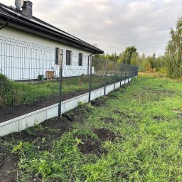 METAL-SIM WOJCIECH SIMLAT - Czarny panelowy system ogrodzeniowy z podmurówką betonową, otaczający posesję z białym domem i zielonym trawnikiem w pochmurny dzień.