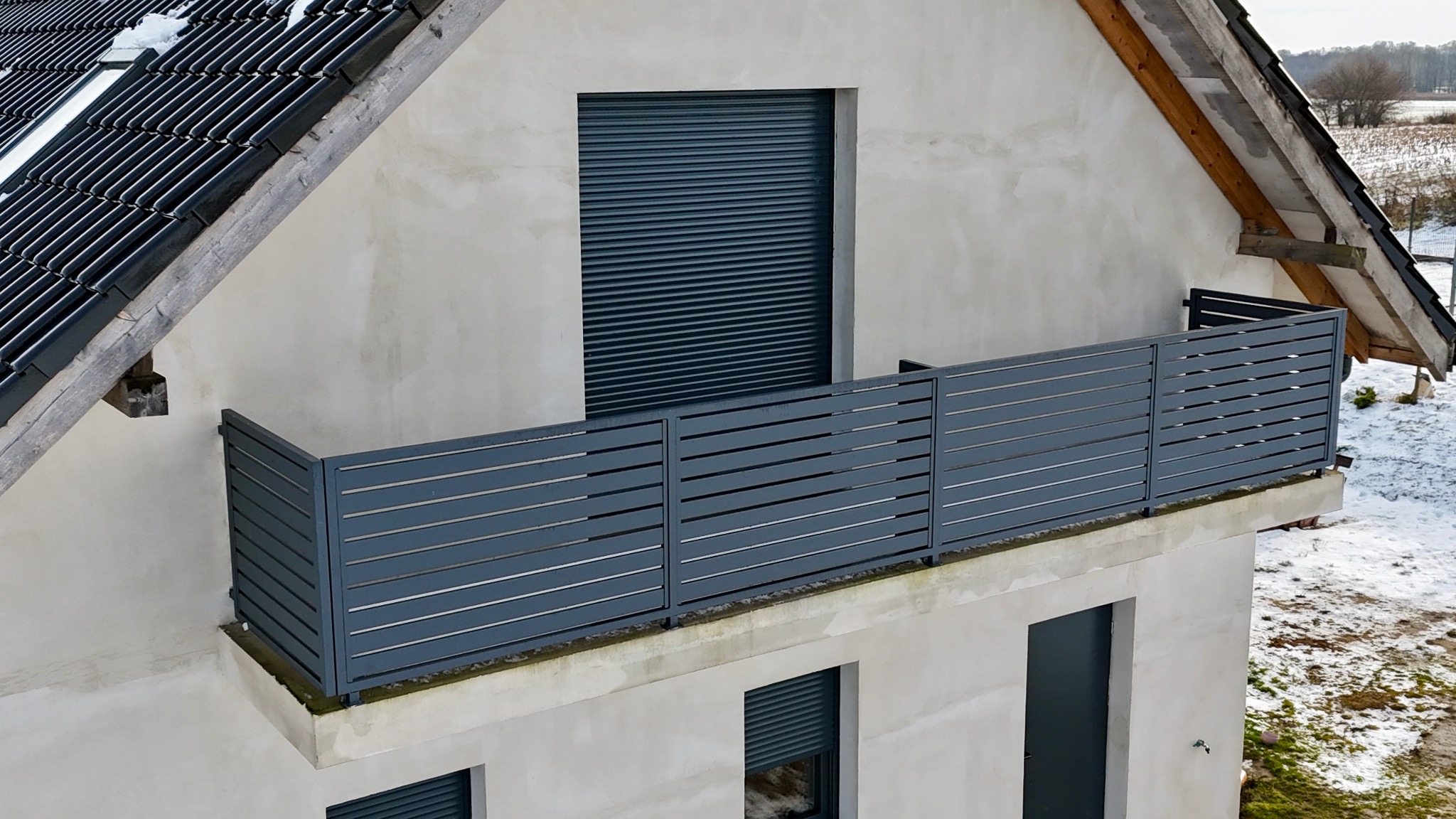 Szara, pozioma balustrada panelowa na balkonie domu z szarą elewacją i roletami zewnętrznymi, widok z góry, zimowy krajobraz w tle.