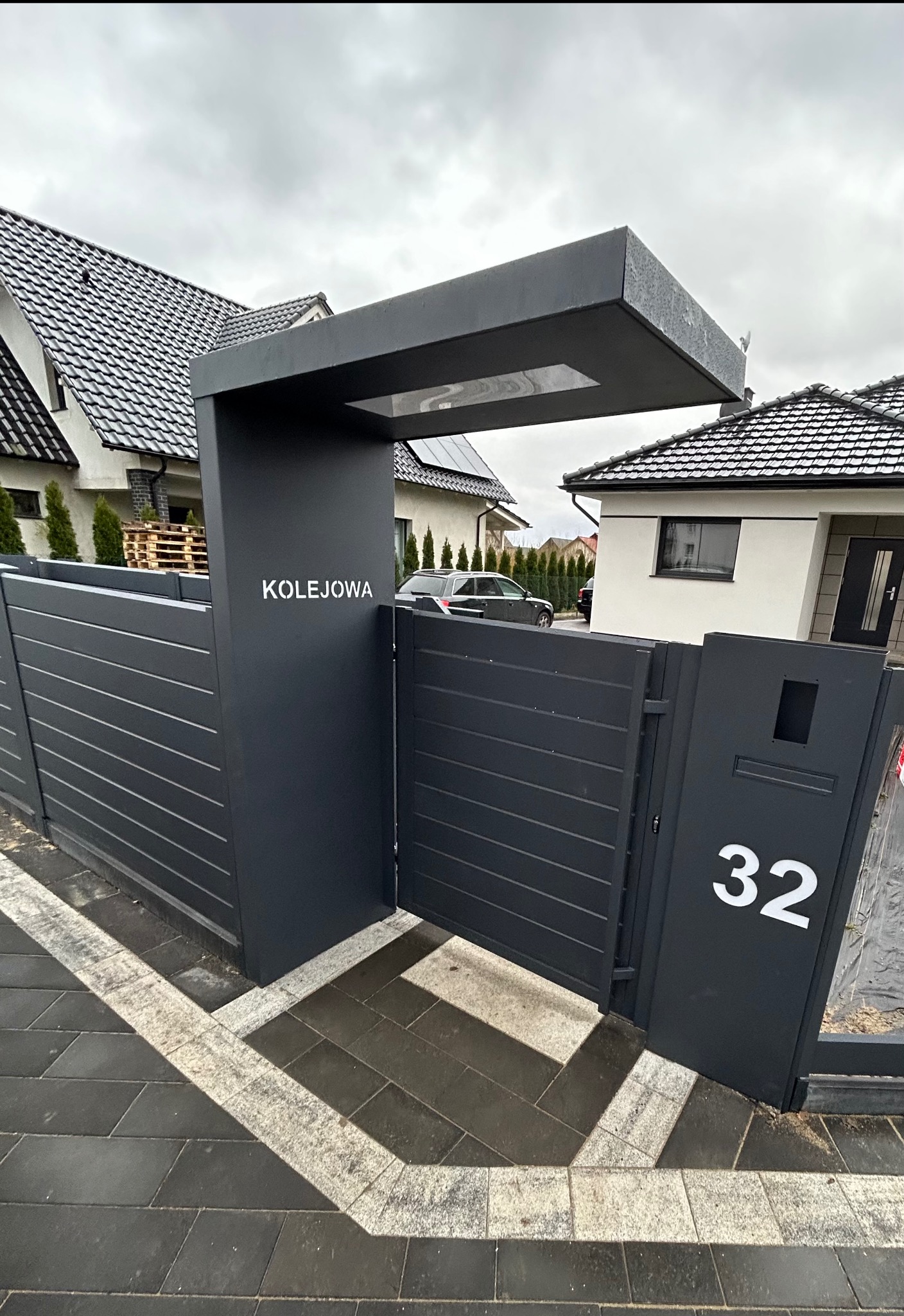 Nowoczesna, grafitowa brama wjazdowa z numerem domu 32 i nazwą ulicy 'Kolejowa', zintegrowana ze skrzynką pocztową, na tle kostki brukowej i domu.
