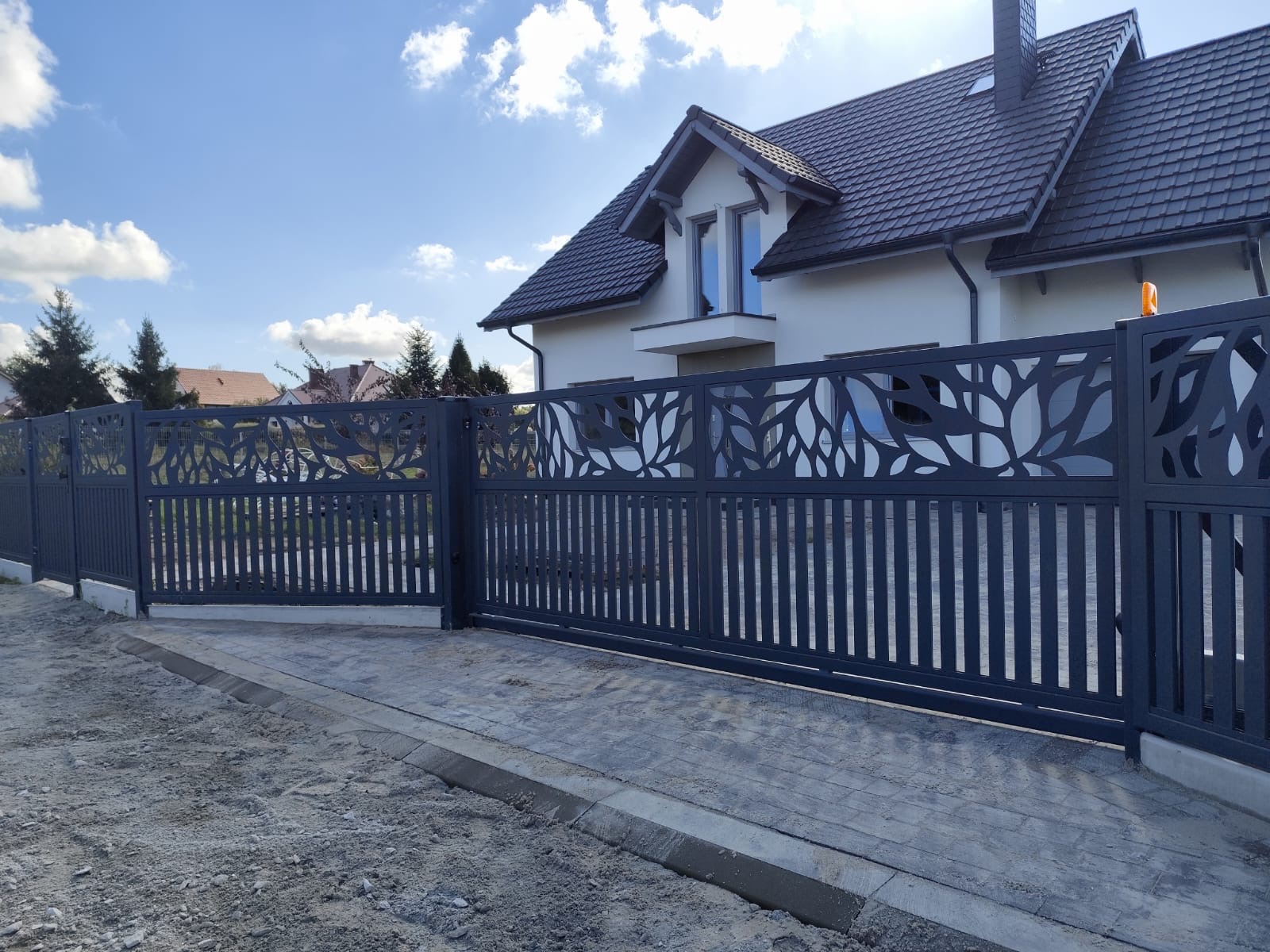 Nowoczesna, przesuwna brama wjazdowa z ażurowym wzorem liści na tle domu z ciemną dachówką.