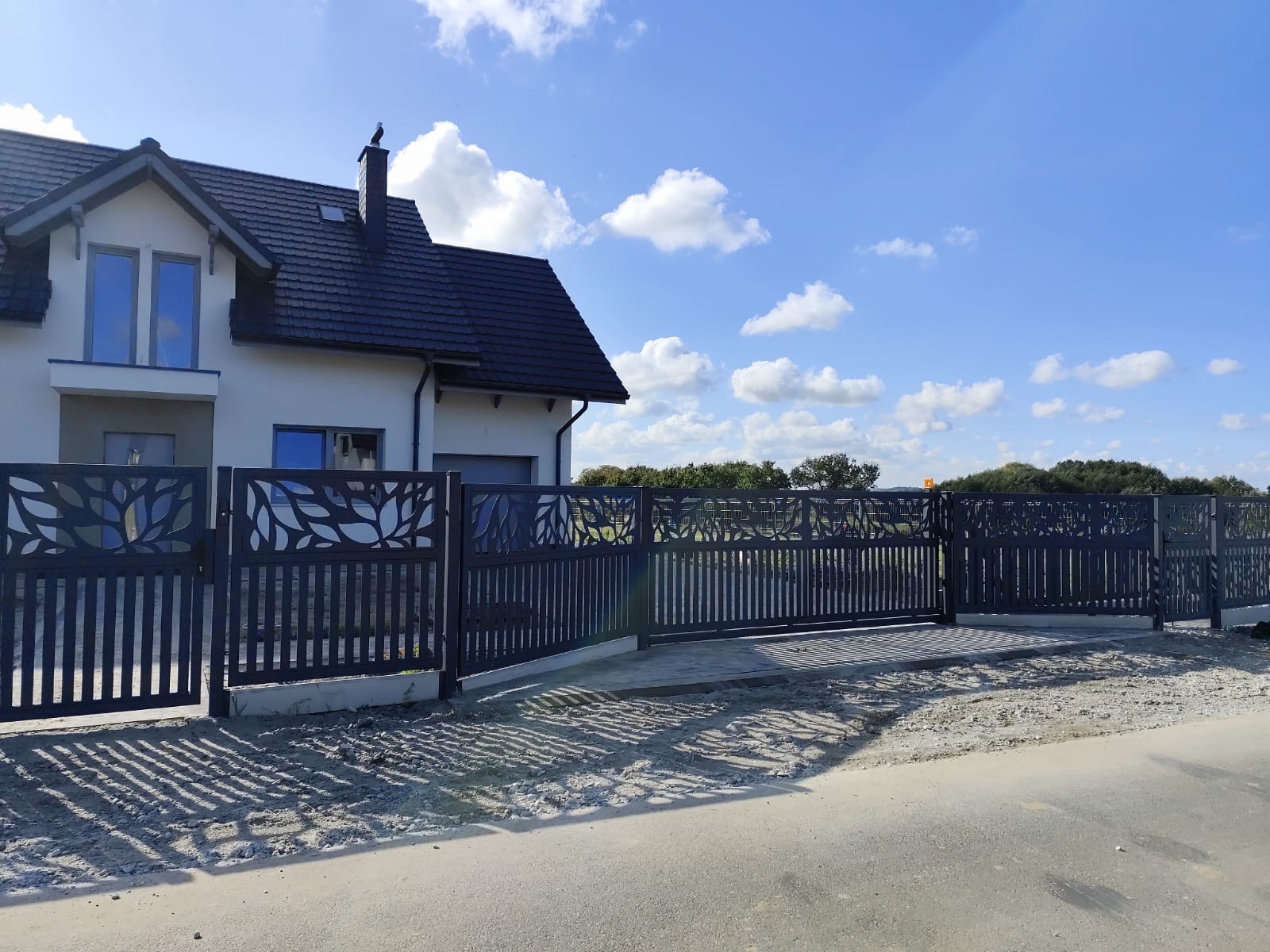 Nowoczesna, metalowa brama wjazdowa z dekoracyjnym motywem liści na tle jasnego domu z ciemnym dachem i błękitnego nieba z chmurami.