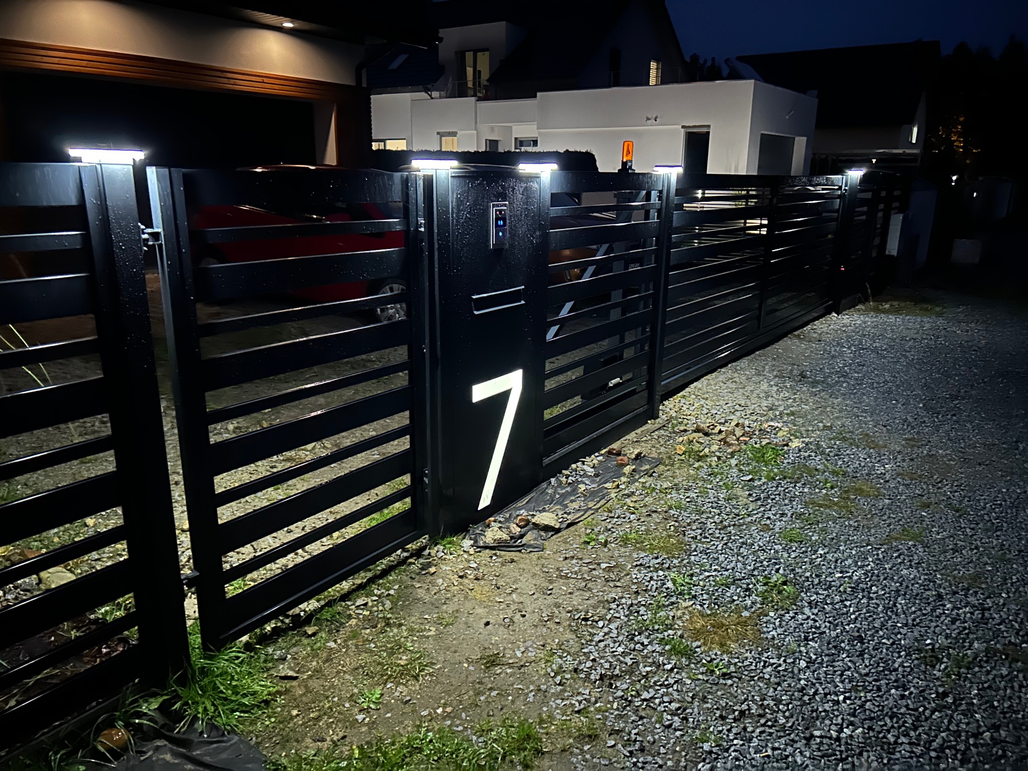 Nowoczesna, czarna brama wjazdowa z numerem domu '7' podświetlonym od wewnątrz, oświetlenie bramy LED, szara nawierzchnia z kamienia na podjeździe, widok na oświetlony dom w tle, nocne ujęcie.