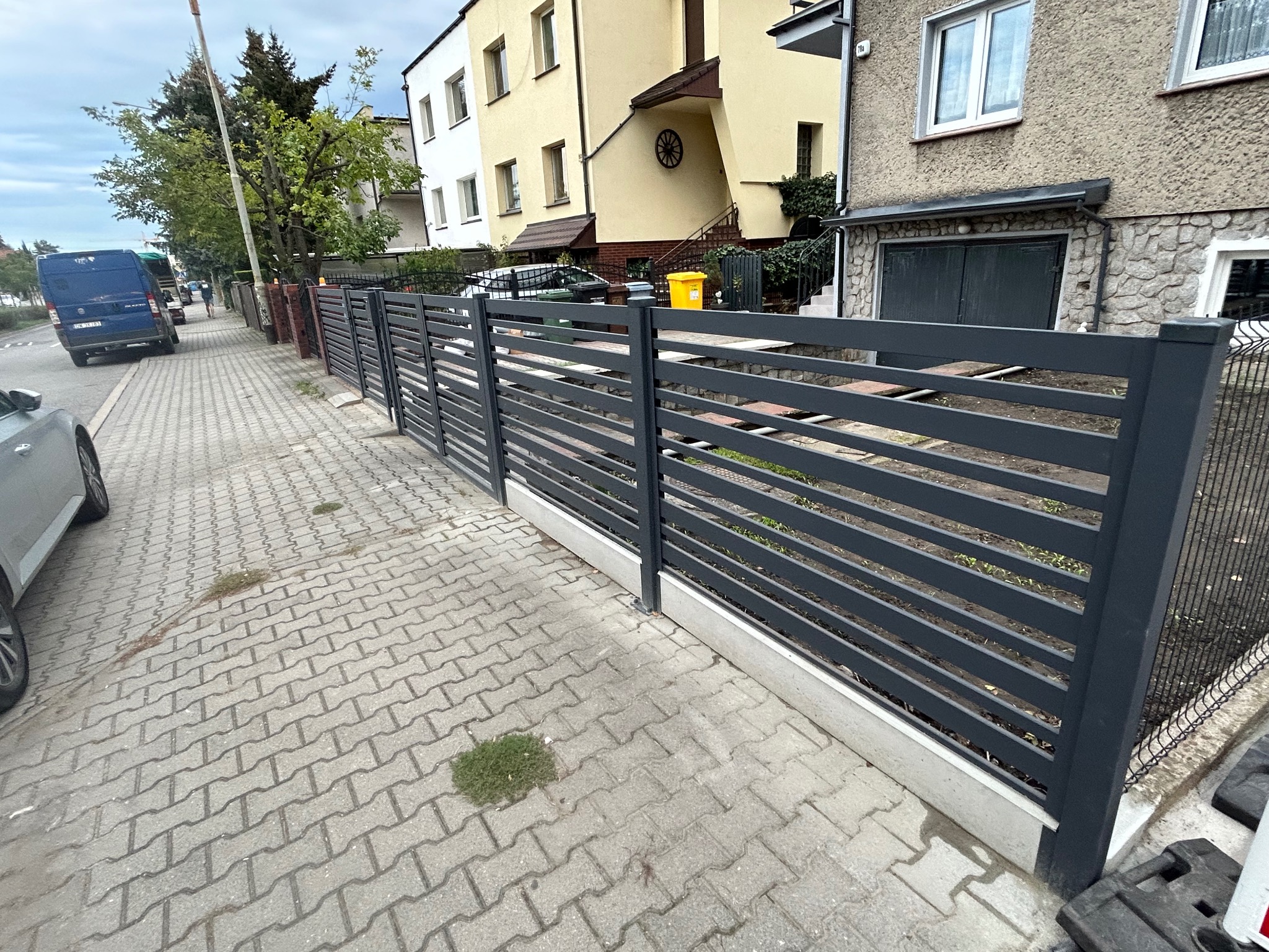 Nowoczesne, poziome przęsła ogrodzenia w kolorze grafitowym, zamontowane na betonowej podmurówce, widok z chodnika z kostki brukowej.