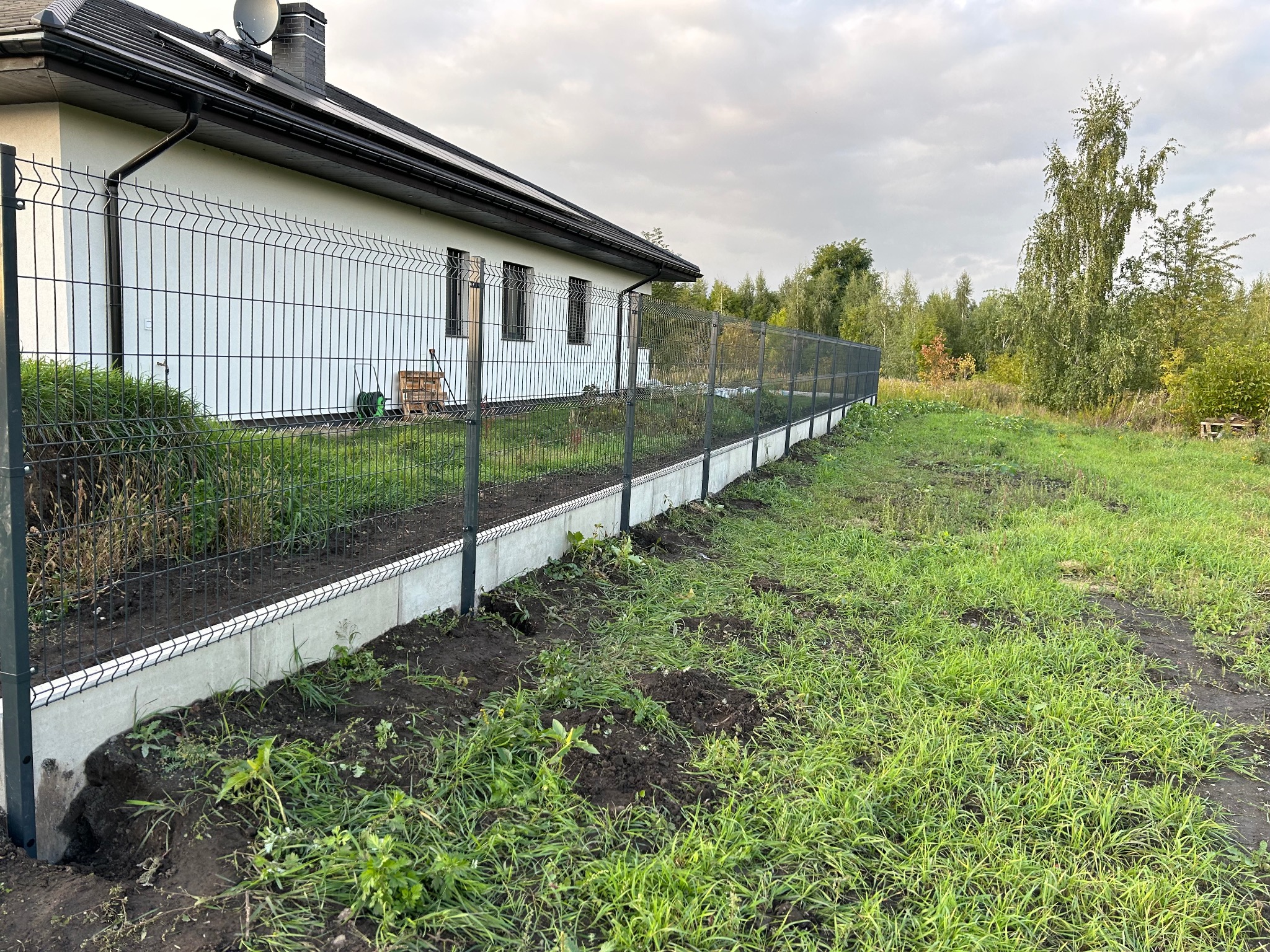 Czarny panelowy system ogrodzeniowy z podmurówką betonową, otaczający posesję z białym domem i zielonym trawnikiem w pochmurny dzień.