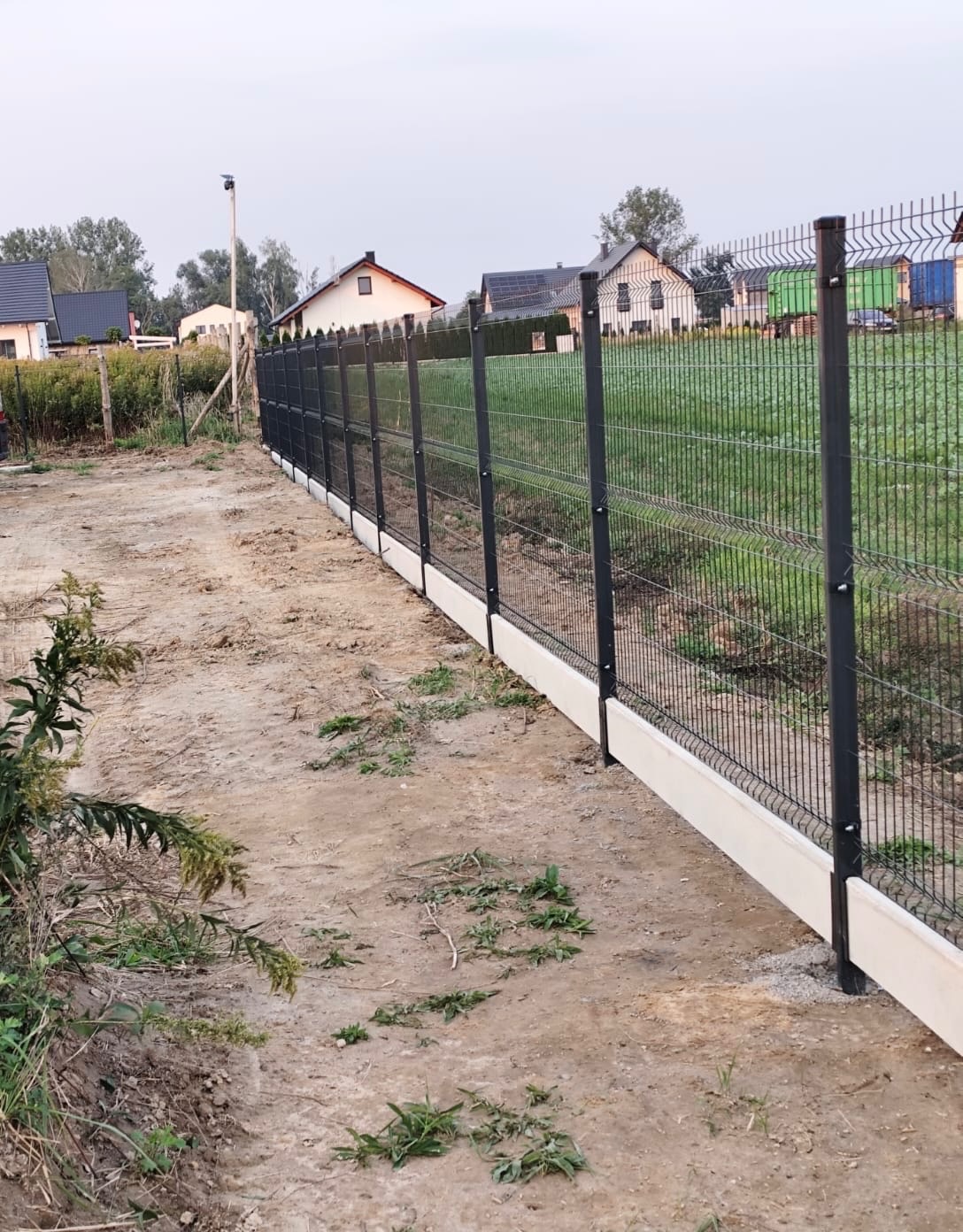 Nowoczesne ogrodzenie z paneli z podmurówką betonową na nierównym terenie, widok z boku.