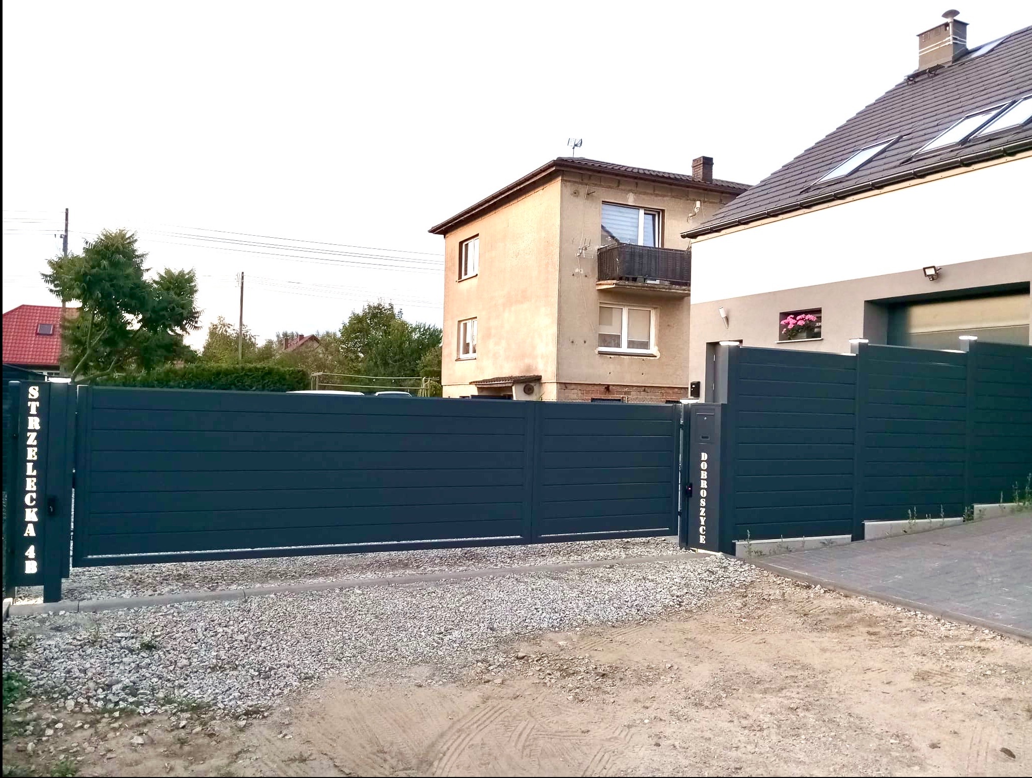 Nowoczesna, pozioma brama wjazdowa i fragment ogrodzenia posesji w Ludgierzowicach, z adresami na słupkach, na tle budynków mieszkalnych.