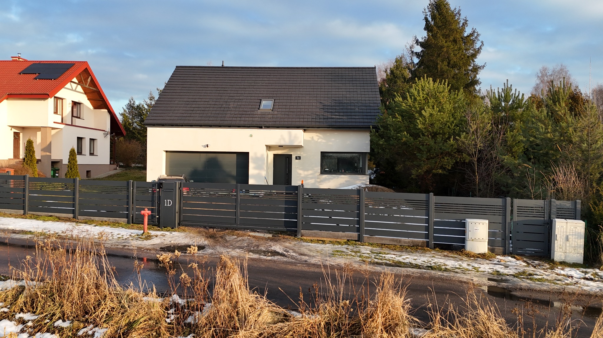 Nowoczesne ogrodzenie panelowe w kolorze grafitowym, z poziomymi szczeblami, otaczające dom z garażem. Widoczne skrzynki gazowe i hydrant. Zimowy krajobraz z resztkami śniegu.