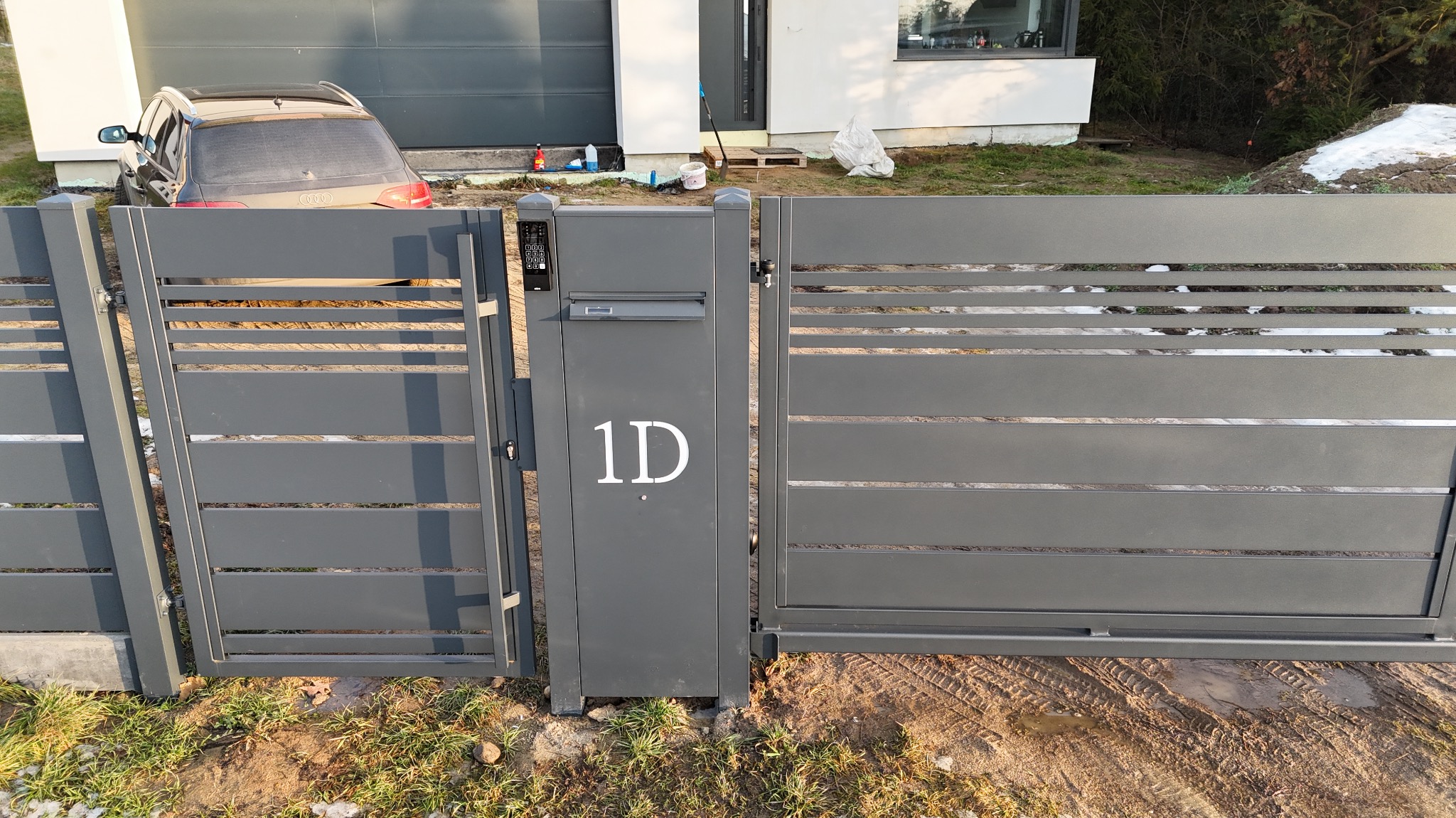 Nowoczesna, pozioma brama wjazdowa z szarego metalu z numerem domu '1D', zintegrowana ze skrzynką na listy i panelem numerycznym, na tle domu i częściowo odśnieżonego trawnika.