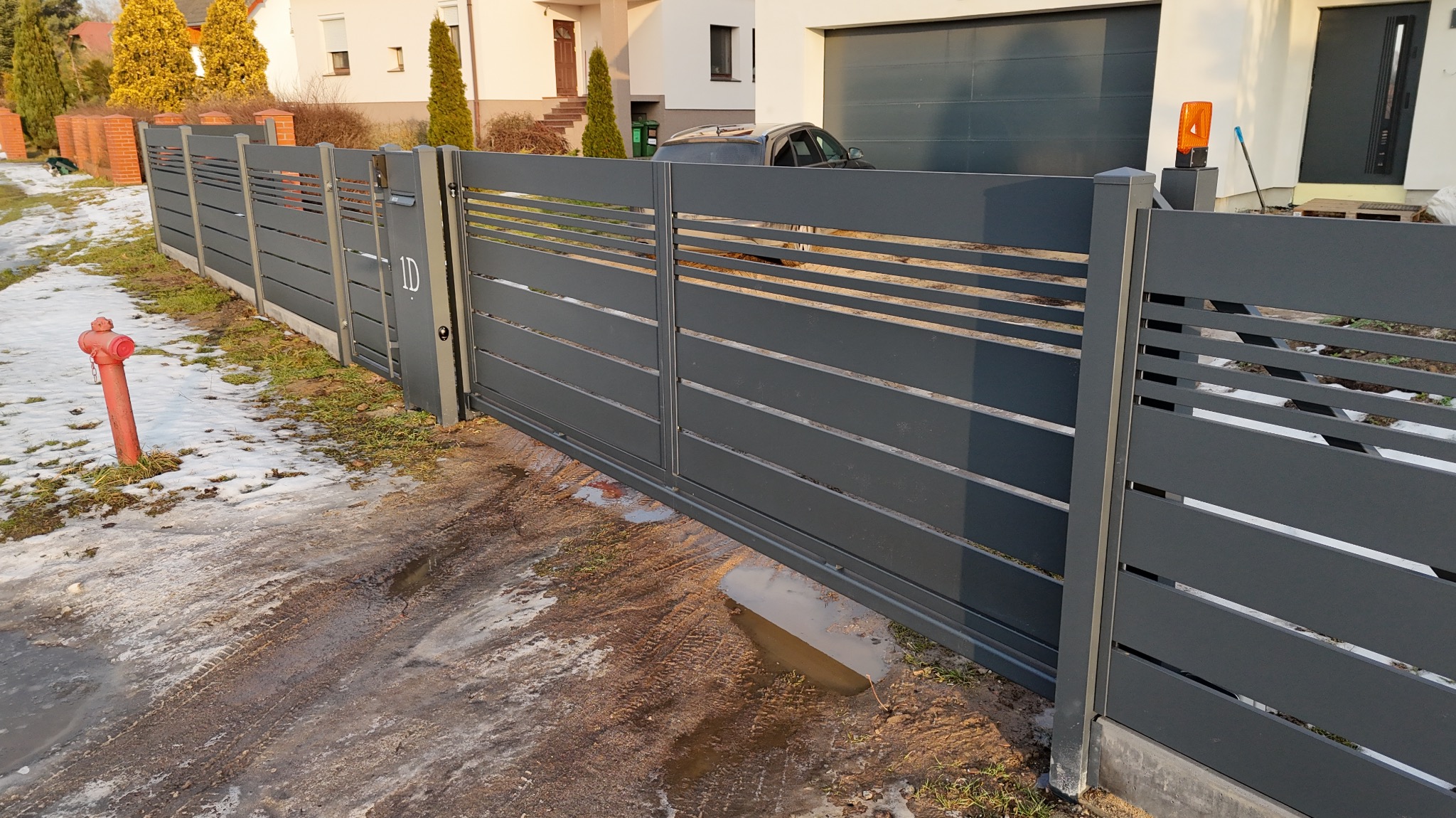 Nowoczesna, przesuwna brama wjazdowa z poziomymi panelami w kolorze grafitowym, z numerem domu '1D', na tle domu z garażem i hydrantu.