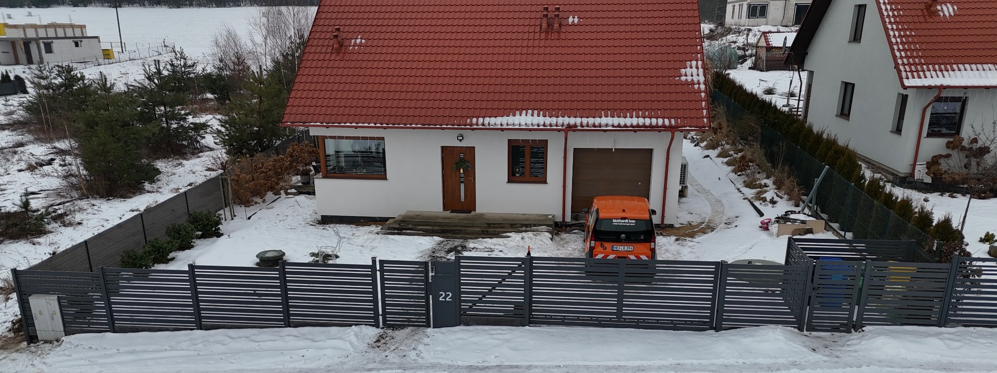 Nowoczesne, poziome ogrodzenie panelowe w kolorze grafitowym, z numerem domu '22', na tle zasypanego śniegiem podwórka i domu z czerwoną dachówką. Widoczny samochód dostawczy.