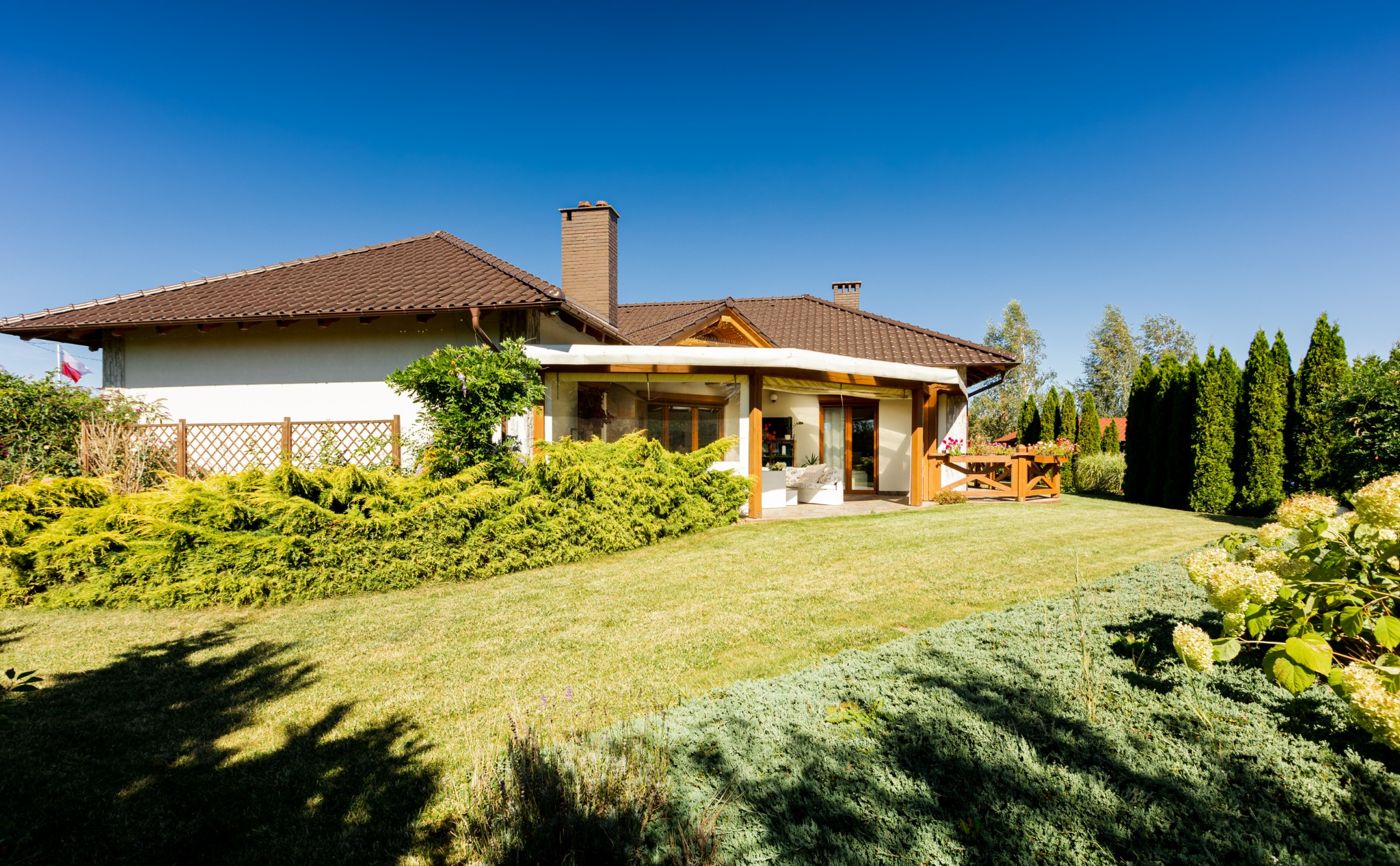 Biały dom z brązowym dachem, widoczny z ogrodu z bujną zielenią i drzewami iglastymi pod błękitnym niebem.