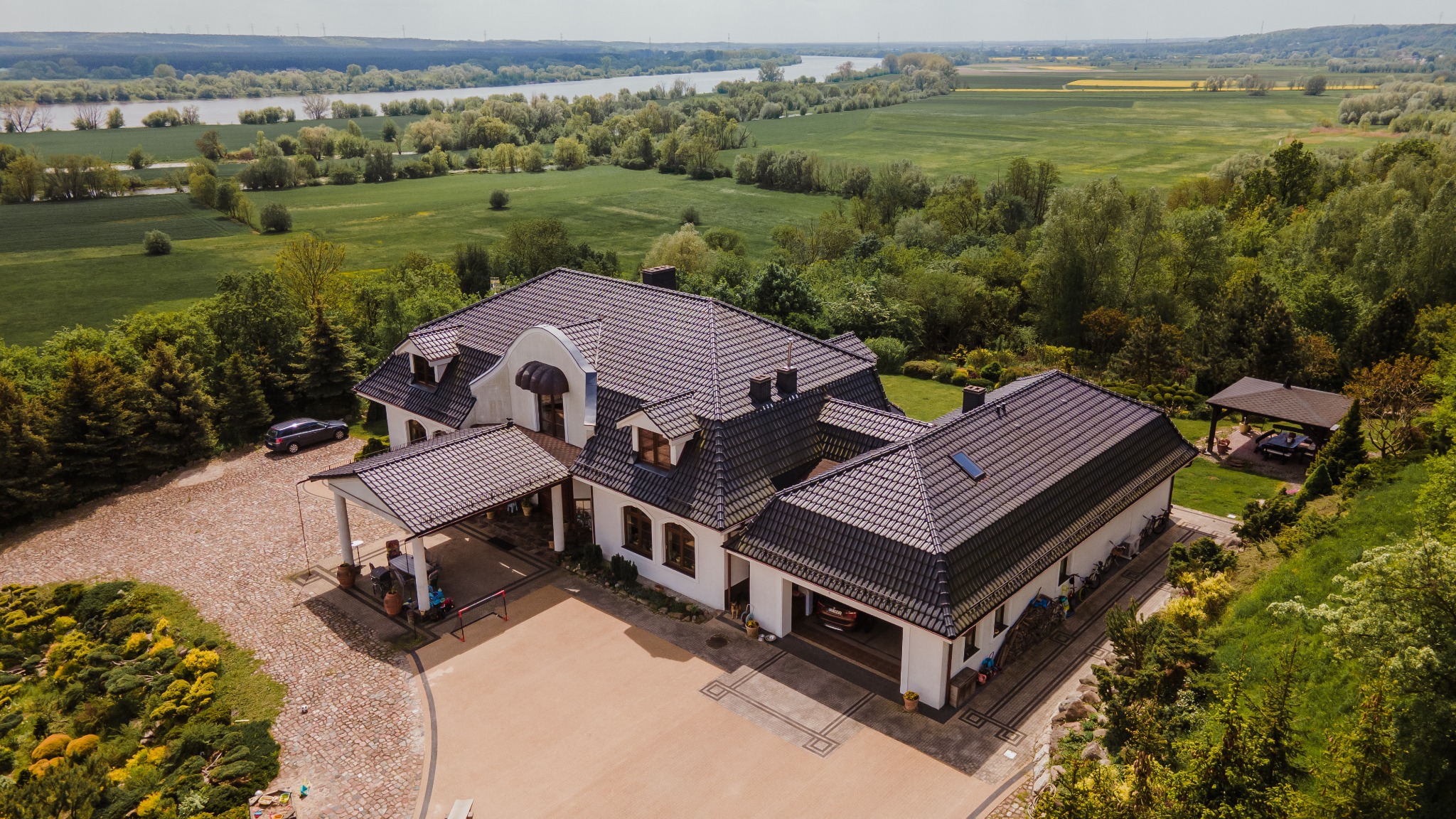 Panoramiczny widok z lotu ptaka na okazałą rezydencję z ciemną dachówką, otoczoną bujną zielenią, z rzeką i polami w tle, sfotografowaną w słoneczny dzień.