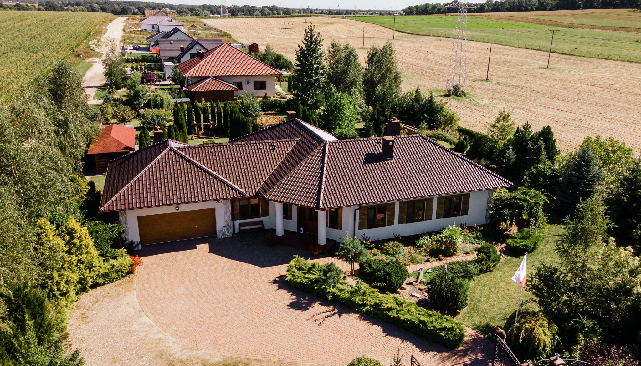 Widok z góry na parterowy dom z brązowym dachem, garażem, brukowanym podjazdem i zadbanym ogrodem z flagą Polski.