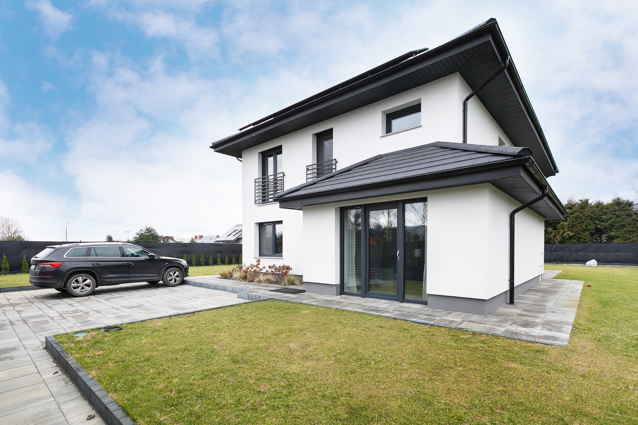 Nowoczesny, dwupiętrowy dom z białymi ścianami i grafitowym dachem, widoczny z boku, z zaparkowanym czarnym SUV-em na wyłożonym kostką podjeździe i zadbanym trawnikiem.