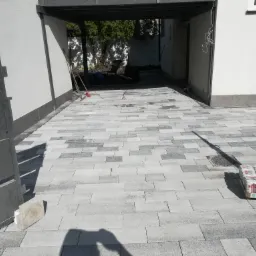 Świeżo ułożona, szara kostka brukowa przed wjazdem do garażu, z paletą napojów w tle i otwartą bramą wjazdową.