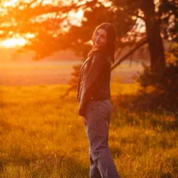 Kobieta w skórzanej kurtce i dżinsach pozuje na łące o zachodzie słońca, odwrócona przez ramię z uśmiechem, w tle drzewo i złote światło.