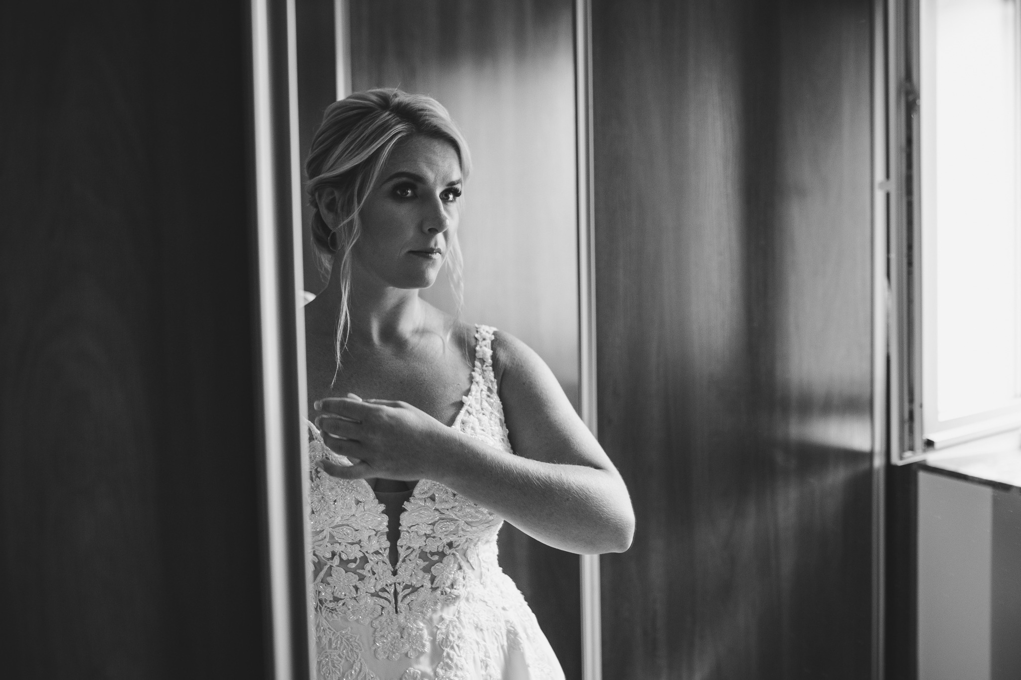 Czarno-białe zdjęcie panny młodej w koronkowej sukni, spoglądającej w bok, w trakcie przygotowań ślubnych przy lustrze.