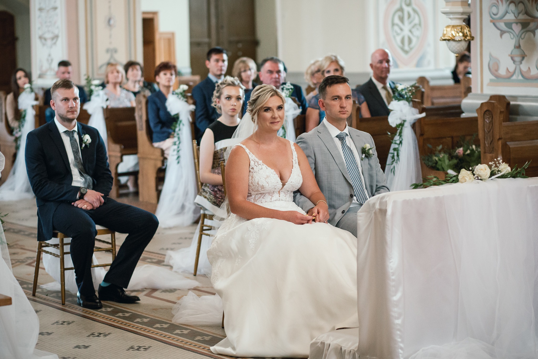 Para młoda siedzi w kościele podczas ceremonii ślubnej, otoczona gośćmi, z dekoracjami kwiatowymi na ławkach. Panna młoda w koronkowej sukni z głębokim dekoltem, pan młody w szarym garniturze.