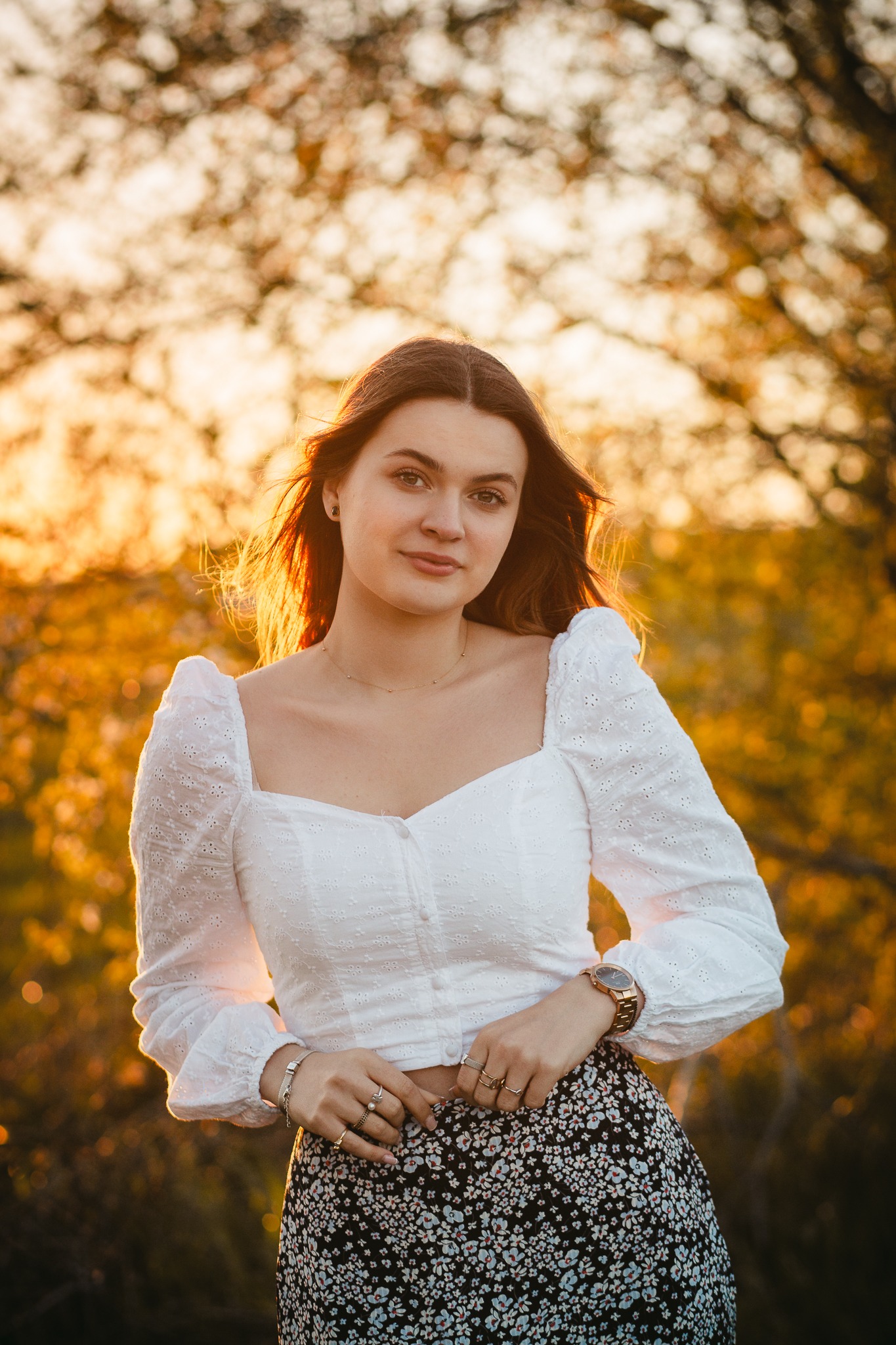 Portret młodej kobiety w białej bluzce z haftem i kwiecistej spódnicy, pozującej na tle drzew w ciepłym, zachodzącym słońcu, z widocznymi pierścionkami i zegarkiem.