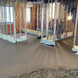 Wnętrze w trakcie prac: świeżo wylana, gładka posadzka, widoczne stalowe profile ścian działowych, izolacja, instalacje elektryczne i fragment siatki zbrojeniowej.