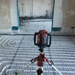 Laser krzyżowy Hilti ustawiony na statywie, widoczny na tle rozłożonego systemu ogrzewania podłogowego przed wylaniem posadzki, z surowymi ścianami w tle.