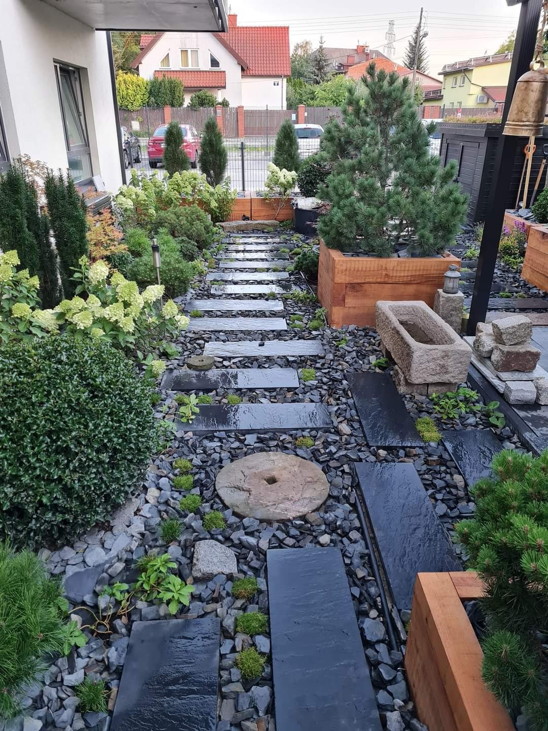 Ogród z ścieżką z ciemnych, prostokątnych płyt kamiennych ułożonych na czarnym żwirze, ozdobiony roślinami, kamienną misą i drewnianymi donicami z iglakami.