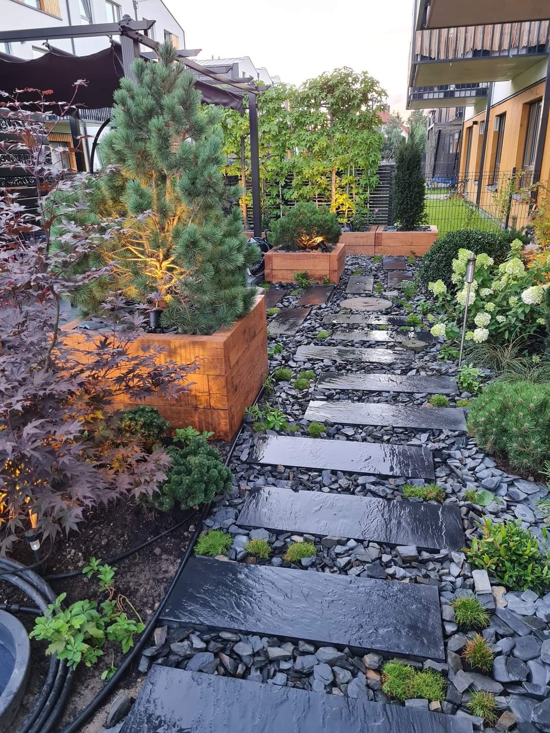 Ścieżka z czarnych płyt kamiennych ułożona wśród drobnych kamieni, oświetlona lampami, z drewnianymi donicami z iglakami i ozdobnymi krzewami w nowoczesnym ogrodzie.