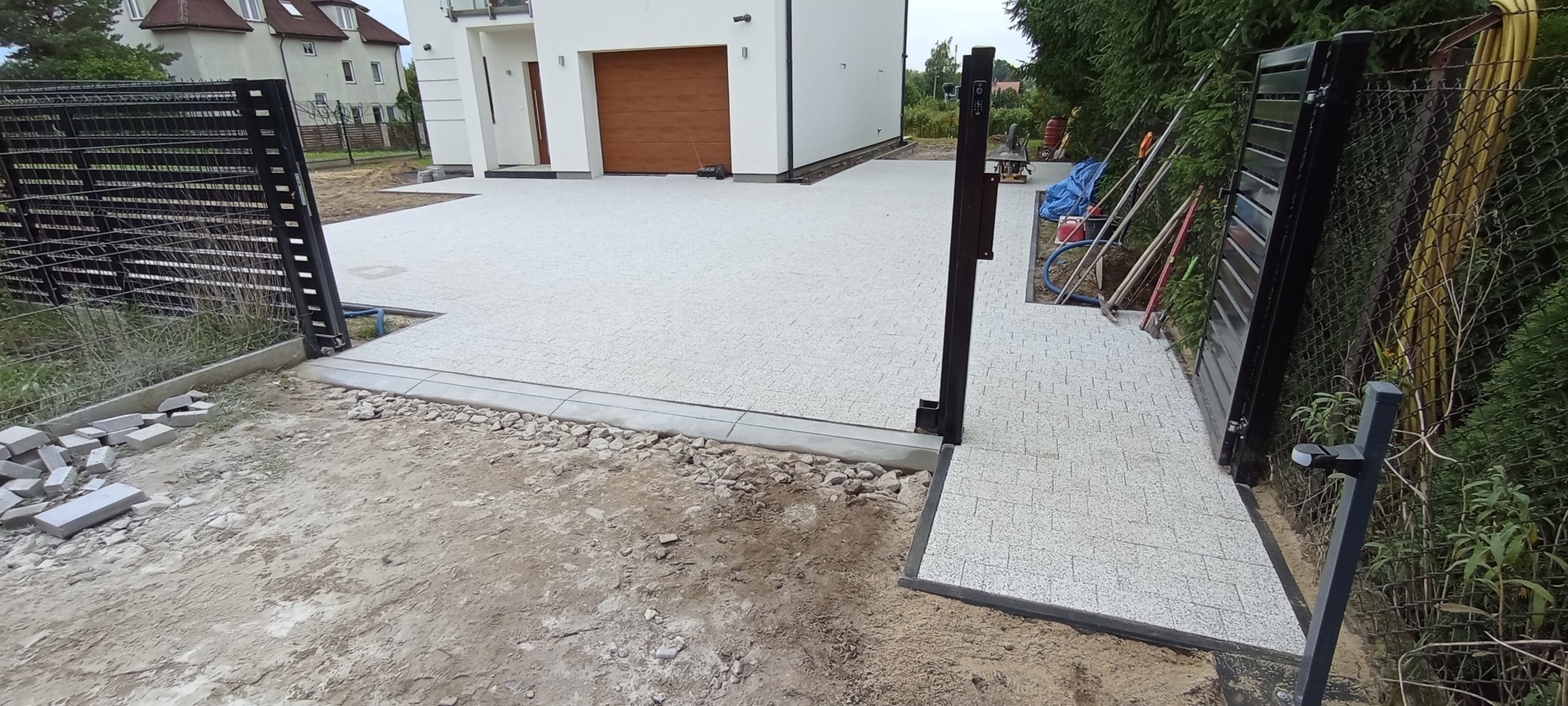 Ułożona jasnoszara kostka brukowa na podjeździe i ścieżce przy nowoczesnym domu z bramą, widoczne narzędzia i materiały budowlane w tle.