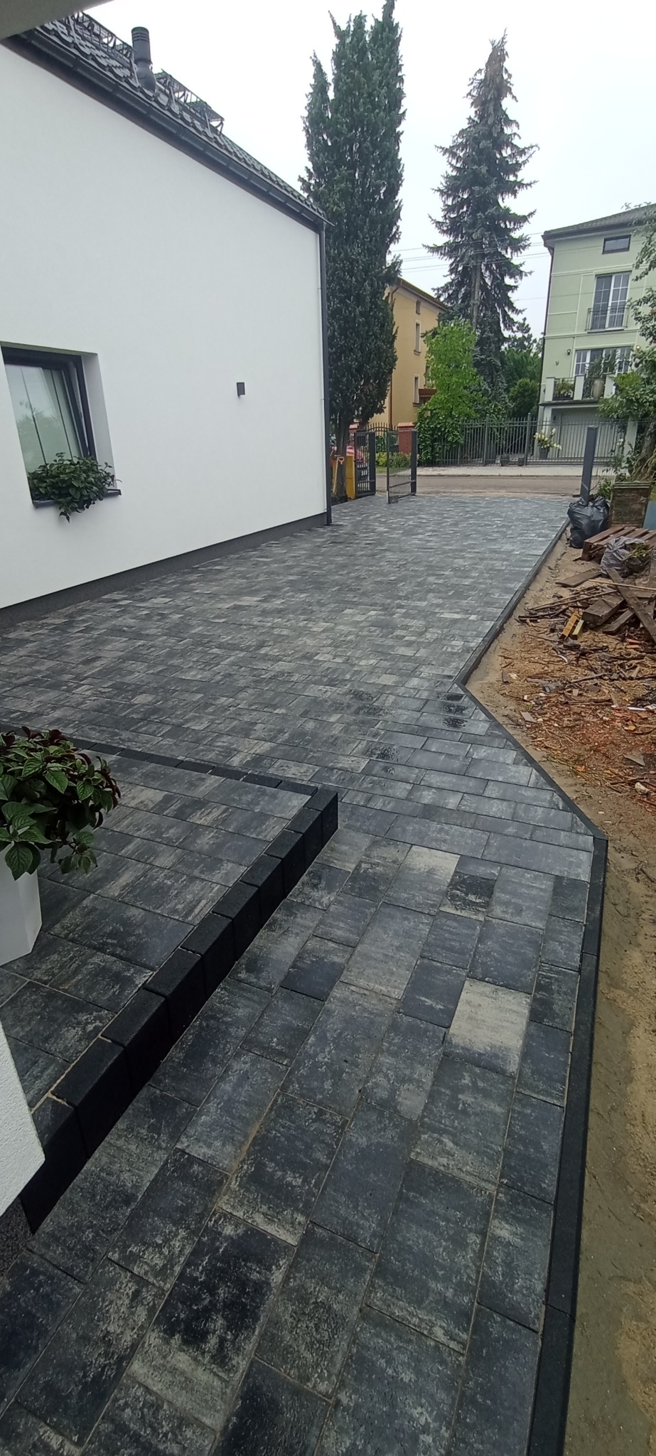 Ukończony podjazd z szarej kostki brukowej prowadzący do domu z białymi ścianami, widoczne rośliny w donicach i fragment ogrodu w tle.