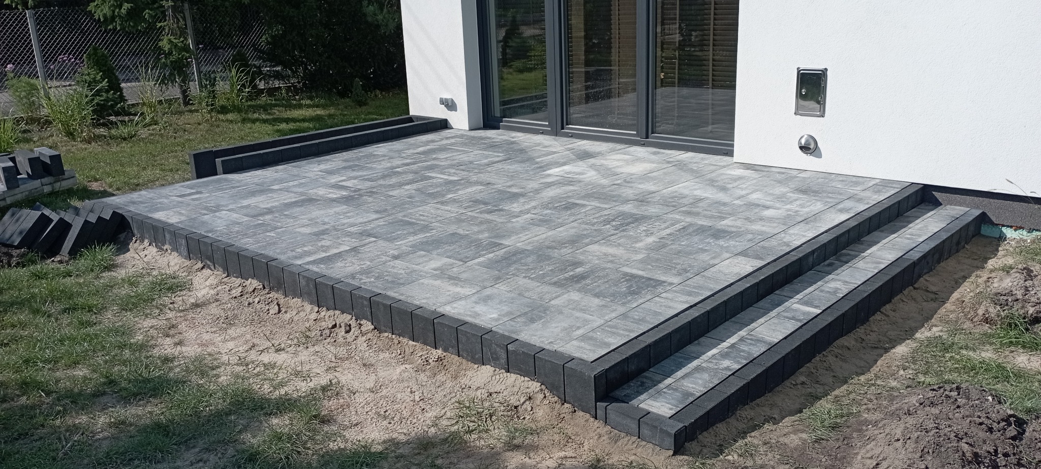 Nowoczesny taras z szarej kostki brukowej z dwustopniowymi schodami, obrzeża z ciemnej kostki, widoczne niedokończone prace ziemne.