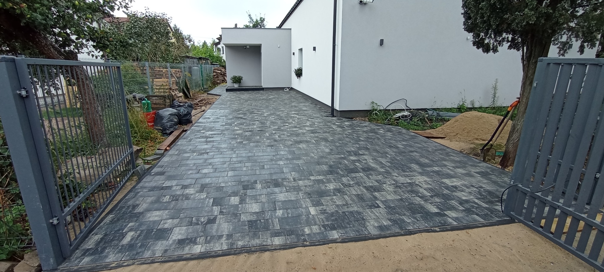 Ułożona, grafitowo-szara kostka brukowa prowadząca do nowoczesnego domu z białymi ścianami i szarym podcieniem, widok zza otwartej, szarej bramy wjazdowej, z widocznym stosem piasku i narzędziami...