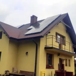 Żółty dom jednorodzinny z ciemnobrązowym dachem, na którym zamontowano panele słoneczne; widoczny komin i balkon z czarną balustradą.