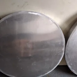 Dwa metalowe, okrągłe elementy, prawdopodobnie po obróbce, leżące na szarym podłożu. Jeden element jest okrągły, drugi ma kształt owalny. Widoczne ślady po szlifowaniu na krawędziach.