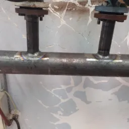 Spawane połączenie rury stalowej z króćcami, widoczne ślady spawania i przebarwienia termiczne, tło stanowi ściana z odpryskującą farbą.