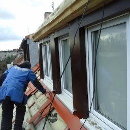 DELA-DACH/SERWIS - Pracownik w niebieskiej kurtce montuje brązowe obróbki blacharskie przy oknach dachowych na pomarańczowym dachu z dachówki ceramicznej, widoczne elementy izolacji i okablowanie.