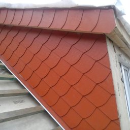 DELA-DACH/SERWIS - Fragment dachu z nowym, czerwonym pokryciem przypominającym łuskę, widoczne elementy konstrukcji dachu i okno.